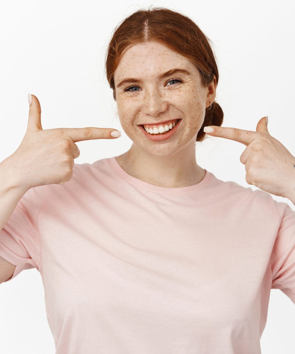 Image of smiling young redhead woman showing her white perfect smile, pointing at whitened teeth without braces, recommending stomatology, dentist advertisement, white background.
